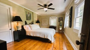 Ein Schlafzimmer mit Holzboden, einem Bett mit weißer Bettwäsche, zwei Nachttischen mit Lampen, einem Deckenventilator und einem Fenster mit Vorhängen.