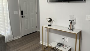 Una sala de estar moderna con un televisor de pantalla plana montado en la pared, una consola blanca con un espejo y una lámpara, y un sofá gris.