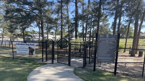 Entrada a un parque canino con un letrero, una puerta metálica y un sendero que conduce al parque.