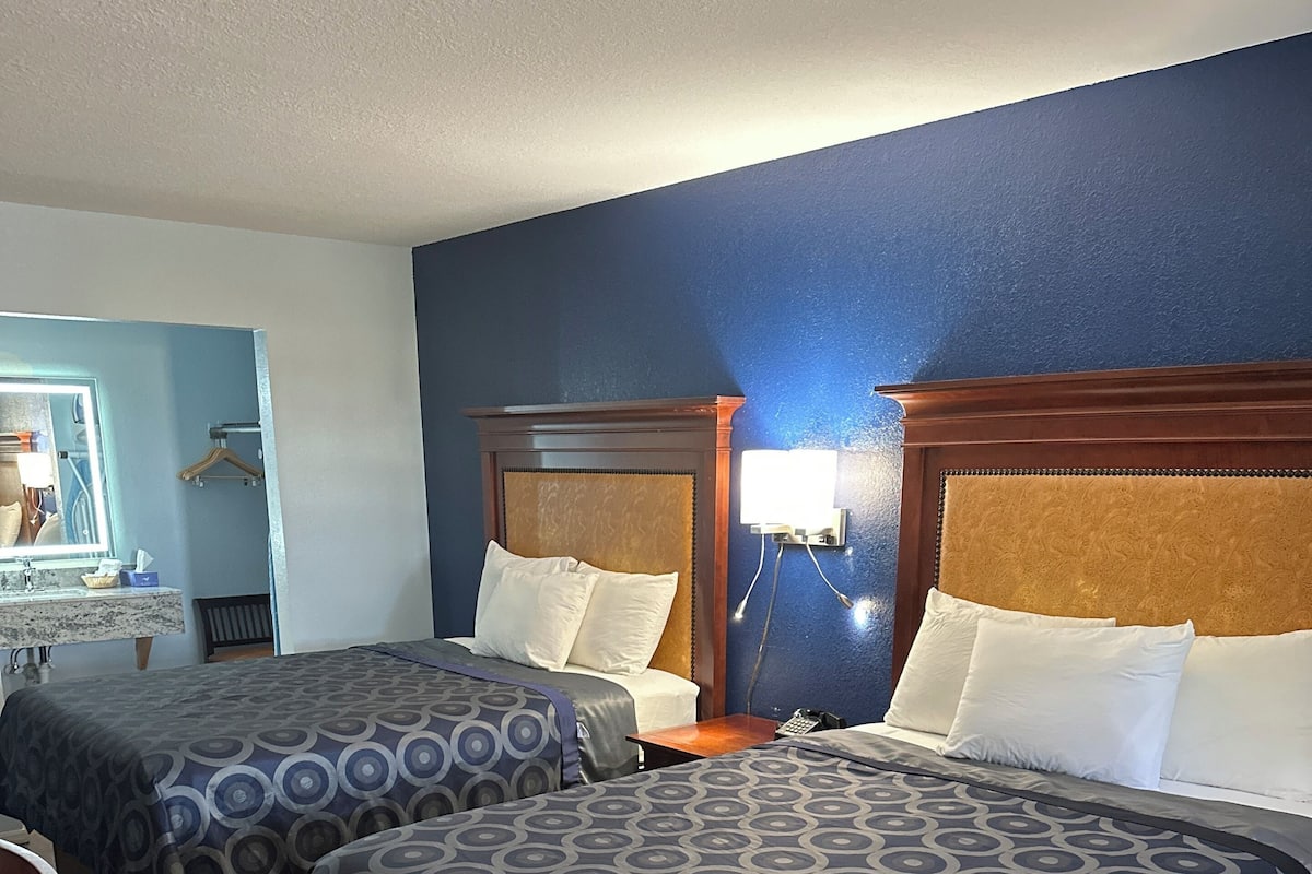 A hotel room with two beds, wooden headboards, a nightstand with a lamp, a mirror, and a wooden chair.