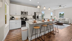 A modern kitchen with white cabinets, stainless steel appliances, a marble island with stools, and a ceiling fan.