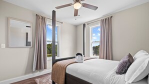 A bedroom with a bed, a ceiling fan, two windows with curtains, and a view of the outdoors.
