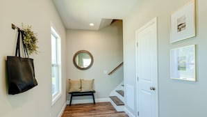 A hallway with a wooden floor, a bench with cushions, a hanging bag, a round mirror, and a staircase with a white door.