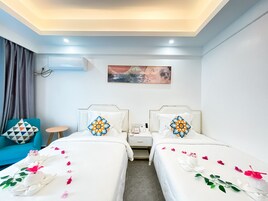 A hotel room with two beds, a small table, a chair, a painting on the wall, and an air conditioning unit.