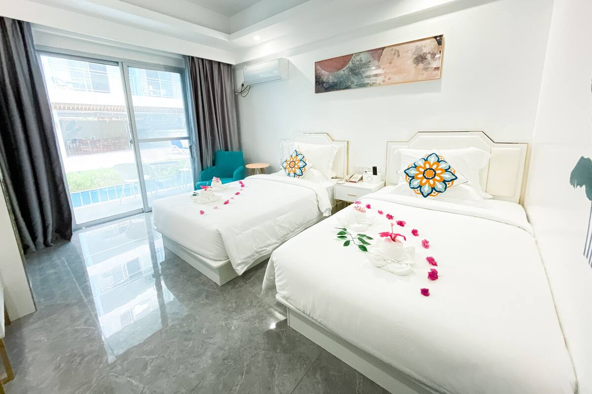 A hotel room with two beds, a large window, a teal chair, and a painting on the wall.