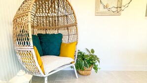 Uma cadeira de vime com almofadas, uma mesinha com um candeeiro e plantas em vasos.