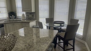 Une cuisine dotée de plans de travail en granit, de meubles blancs et d’un coin repas avec une table et des chaises en verre.