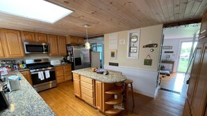A kitchen with wooden cabinets, a granite countertop, stainless steel appliances, and a central island with stools. - Classic Cape Steps from the Beach (Barnstable)