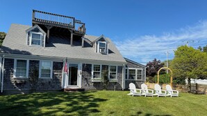 A two-story house with a covered porch, a small deck, and a fenced yard with Adirondack chairs. - Classic Cape Steps from the Beach (Barnstable)