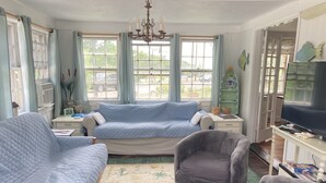 A cozy living room with a blue sofa, a grey armchair, and a matching ottoman. There is a television on a stand, a bookshelf, and a window with curtains. - Classic Cape Steps from the Beach (Barnstable)