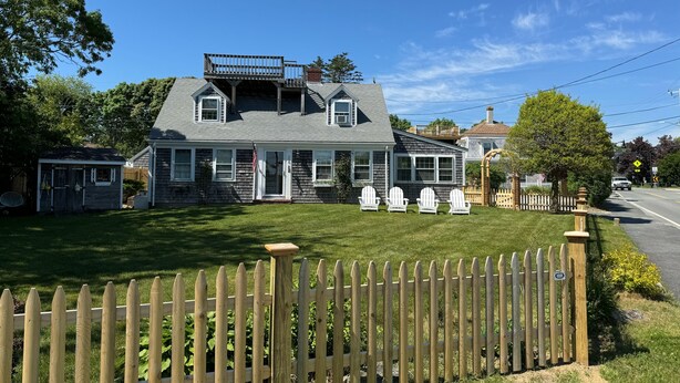 A two-story house with a front porch, a deck, and a fenced yard. - Classic Cape Steps from the Beach (Barnstable)