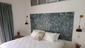 Una cama cuidadosamente tendida con sábanas blancas y almohadones decorativos, un gran cuadro de textura en la pared encima, y dos mesitas de noche con lámparas y pequeñas macetas.
