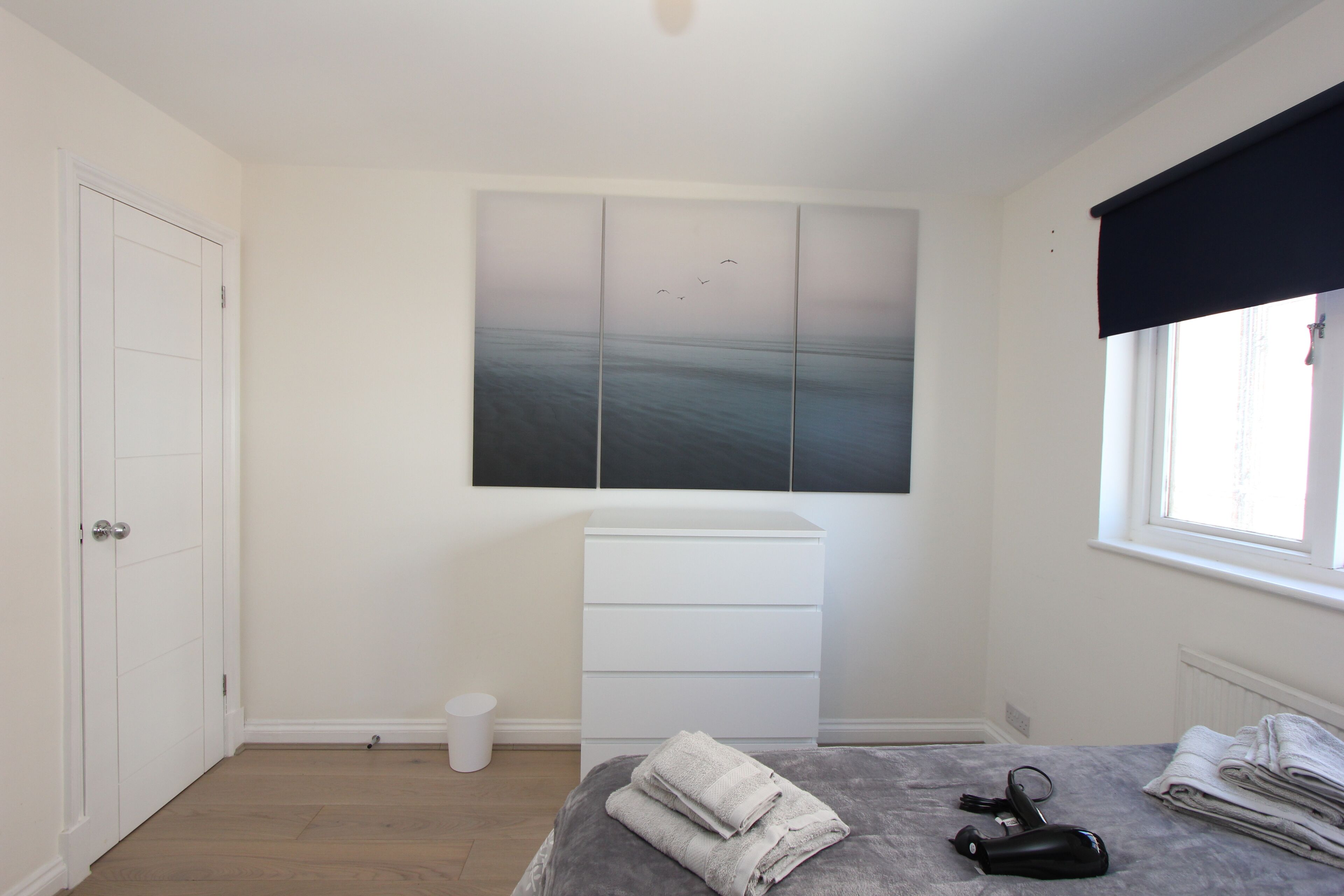 A bedroom with a bed, a dresser, a window with a blackout blind, and a painting on the wall.