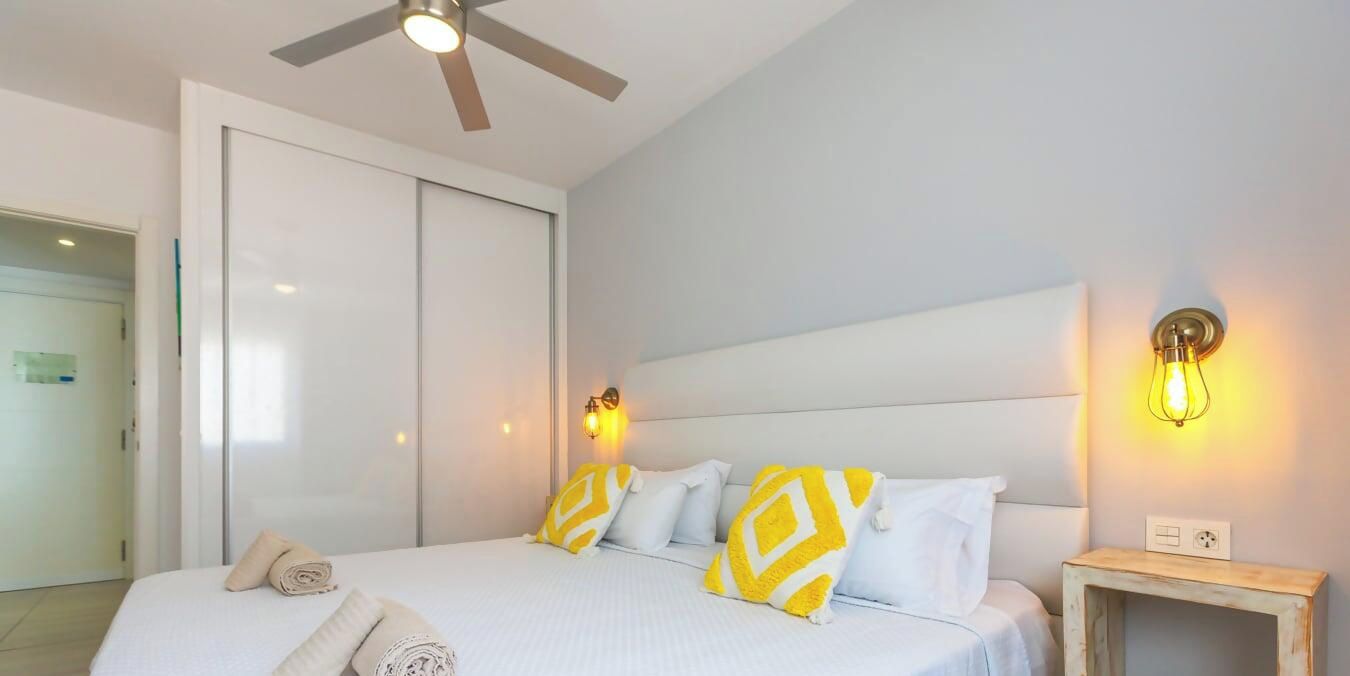 A hotel room with a bed, a ceiling fan, a bedside table, and a wall-mounted light fixture.