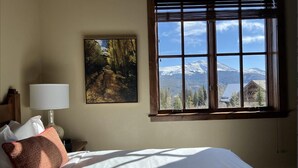 Een slaapkamer met een groot bed, een nachtkastje met een lamp, een schilderij van een bos en een raam met uitzicht op bergen.