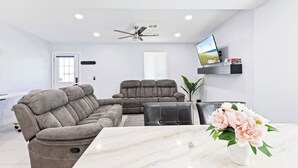 Un salon moderne avec un canapé d’angle, un fauteuil inclinable, une table basse, un téléviseur fixé au mur et une plante.