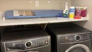 A laundry area with two front-loading washing machines, a shelf with cleaning supplies, and a blue ironing board.