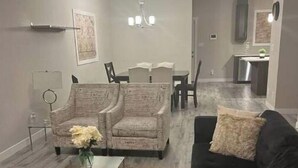 A modern living room with a black sofa, patterned armchairs, a glass coffee table, and a dining area in the background.