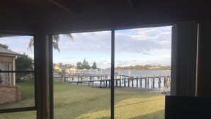 A view through large windows of a grassy area with a dock extending into a body of water, surrounded by buildings and trees. - 3 Bedroom house, right on the river, own private jetty (Greenwell Point)