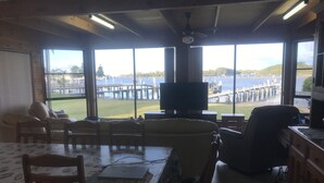 A living room with a large window overlooking a dock and water, a television, and a dining area with a table and chairs. - 3 Bedroom house, right on the river, own private jetty (Greenwell Point)