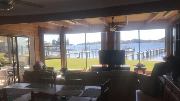 A spacious room with large windows offering a view of a dock and water. There is a dining table with chairs and a television on a stand. - 3 Bedroom house, right on the river, own private jetty (Greenwell Point)