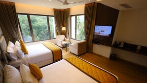Habitación de hotel con dos camas, televisión, una silla y ventanas amplias con vistas al verdor.