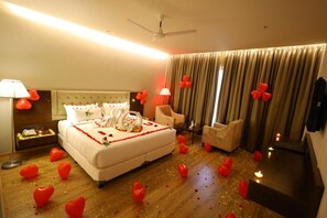 A hotel room with a bed, a nightstand, a chair, a TV, and a ceiling fan. - The Aurum Subrahmanya (Sullia)