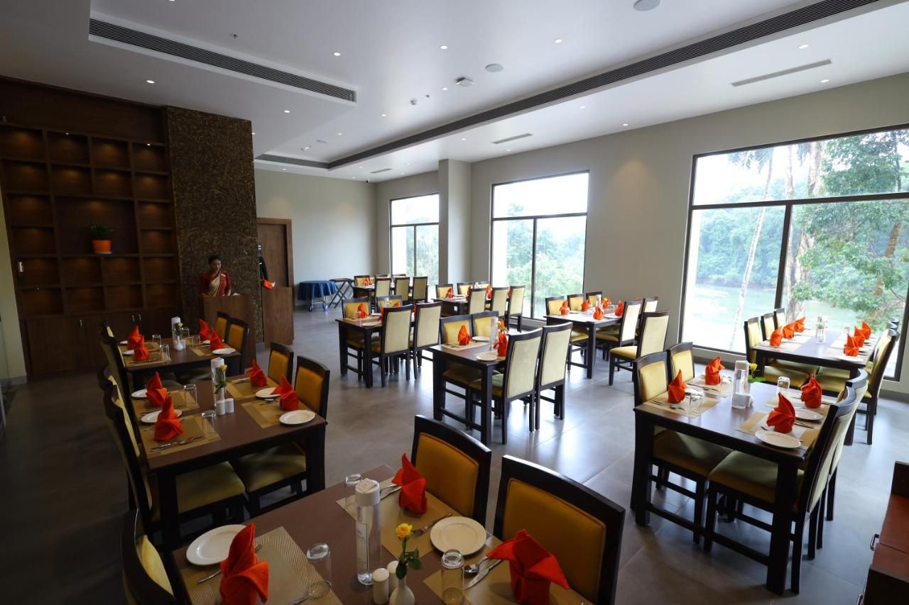 A spacious dining area with multiple tables set for guests, large windows offering a view of the outdoors, and a reception area with a person standing near the entrance.