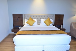 A hotel room with a large bed, two nightstands, a wooden headboard, and a brown armchair. - The Aurum Subrahmanya (Sullia)