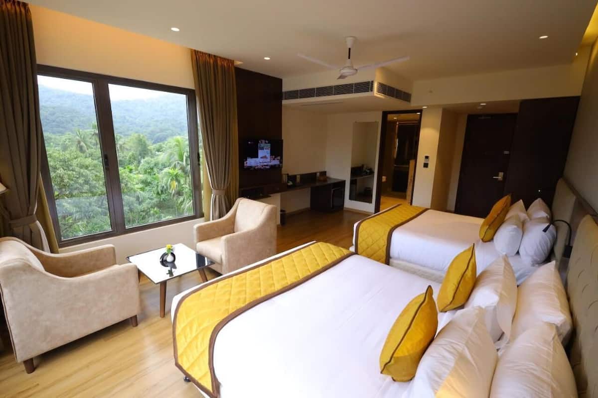 A hotel room with two beds, a sofa, a TV, and a balcony with a view of greenery.
