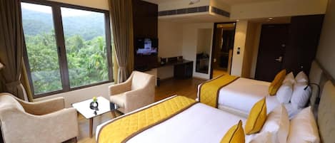 A hotel room with two beds, a sofa, a TV, and a balcony with a view of greenery.