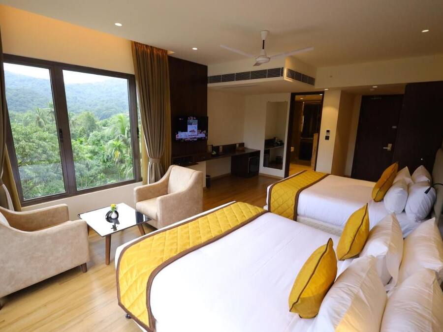 A hotel room with two beds, a sofa, a TV, and a balcony with a view of greenery.