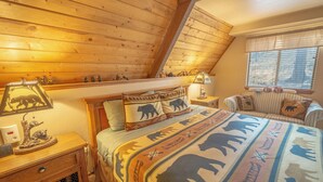 A cozy room with a sloped ceiling, a bed with a bear-themed blanket, a bedside table with a lamp, and a window with blinds. - Cozy Upper Canyon Gem w Large deck and Great Forest Views (Ruidoso)