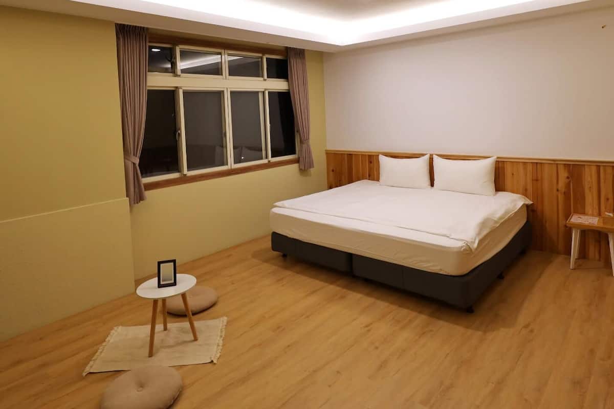 A bedroom with a bed, a window with curtains, a small table with a picture frame, and a bedside table.