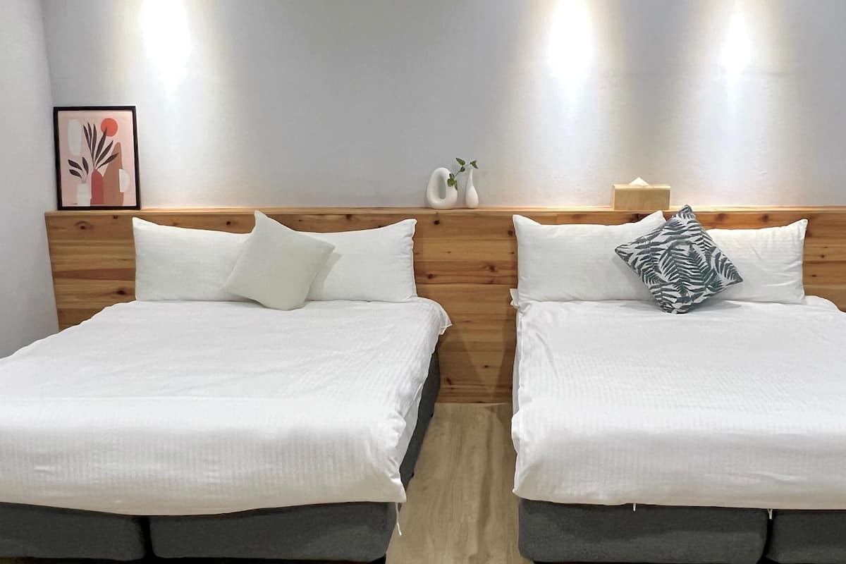 A hotel room with two beds, each with white bedding and a wooden headboard. There are wall-mounted lights above the beds and a framed picture on the wall.