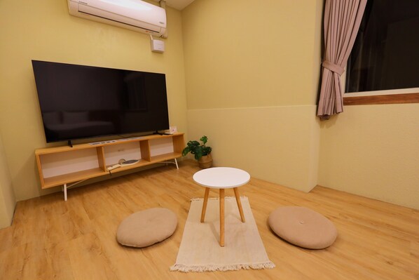 Une pièce avec un téléviseur à écran plat posé sur un meuble en bois, une petite table ronde avec un tapis blanc, deux fauteuils en forme de poire, une plante en pot et une fenêtre avec des rideaux.