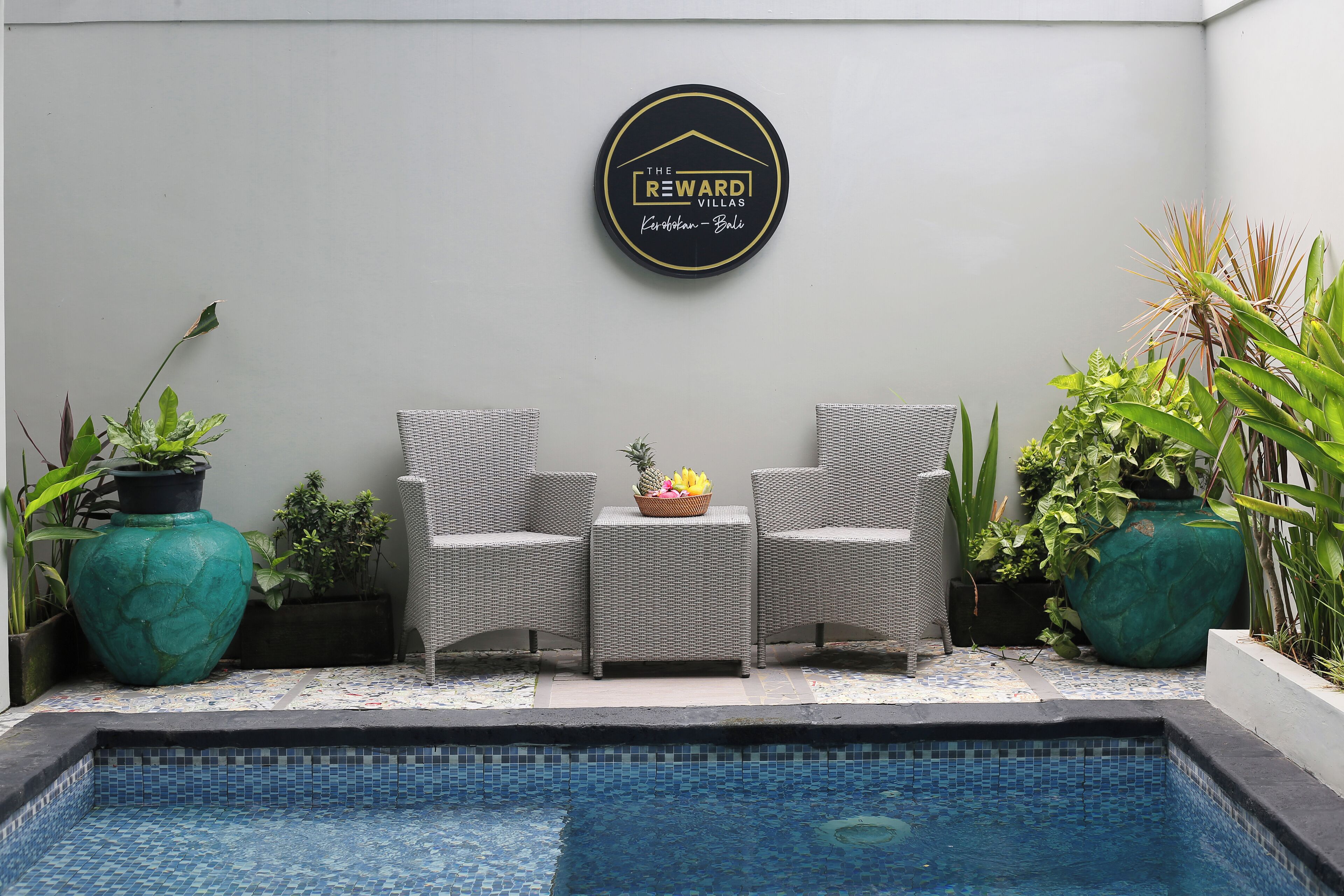 Un espace piscine agrémenté de deux fauteuils en osier, d’une table sur laquelle est posé un panier de fruits, et d’un panneau indiquant « The Reward Villas ».