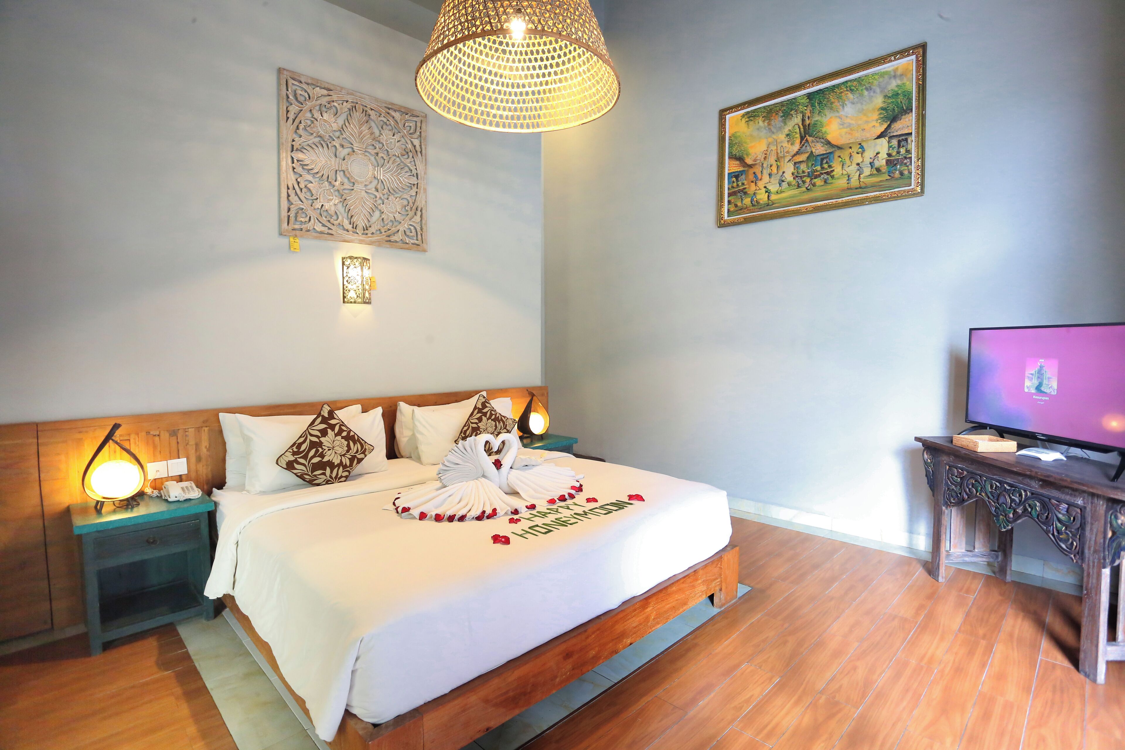 A neatly made bed with a swan decoration and rose petals, a television on a wooden stand, and a painting on the wall.