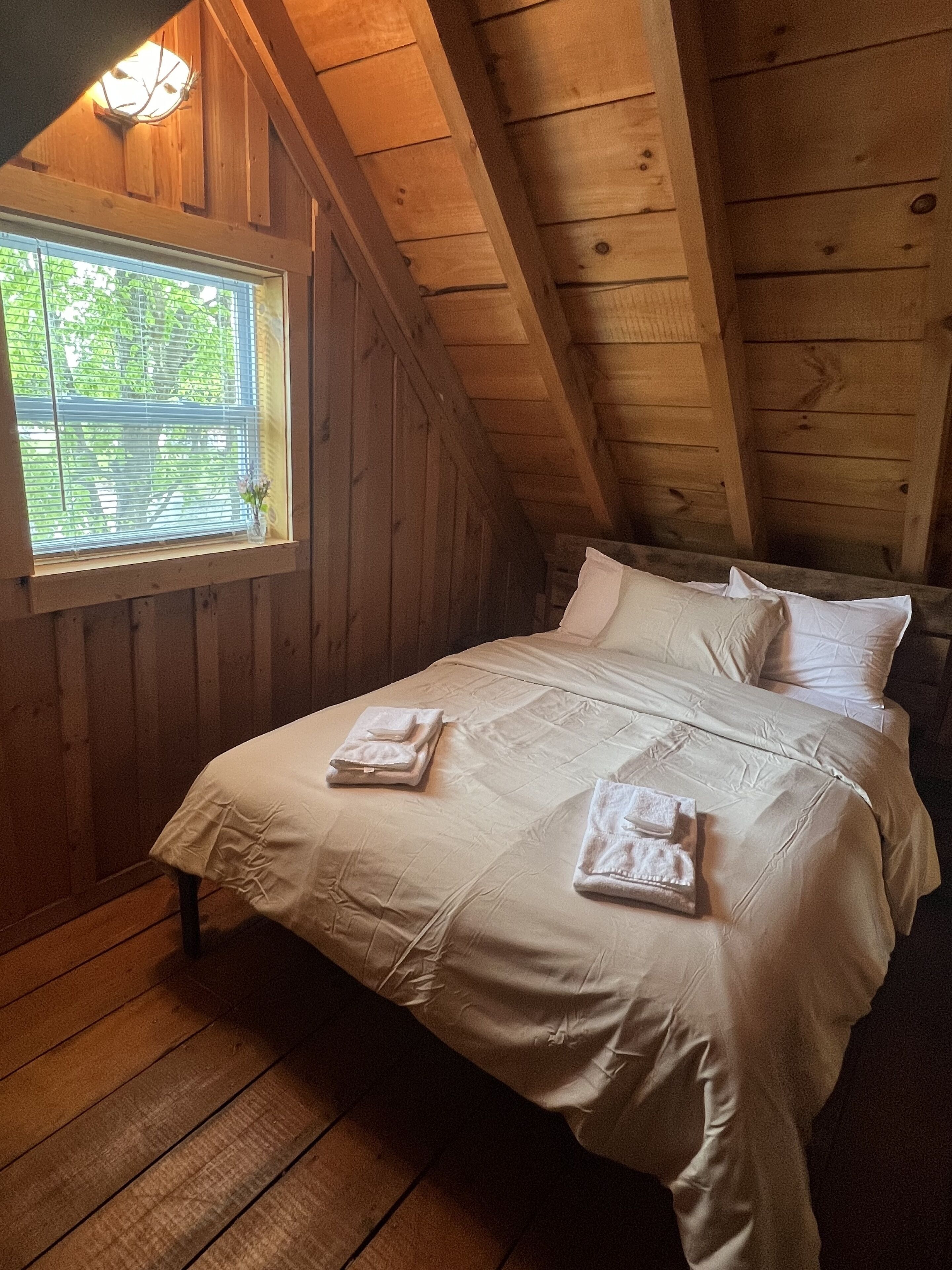 Une pièce avec un plafond en bois, un lit recouvert de draps blancs et une fenêtre équipée de stores.