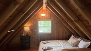 A cozy attic bedroom with a wooden sloped ceiling, a bed with white linens, a nightstand with a lamp, and a window with a view of greenery.