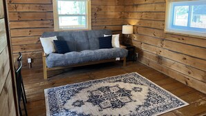 A cozy cabin room with a wooden interior, a sofa, a patterned rug, and two windows.