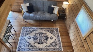 A wooden cabin interior with a gray sofa, two black cushions, a patterned rug, and a window with blinds.