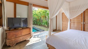 Een slaapkamer met een bed, een televisie op een houten dressoir en uitzicht op een zwembad en tropische planten.