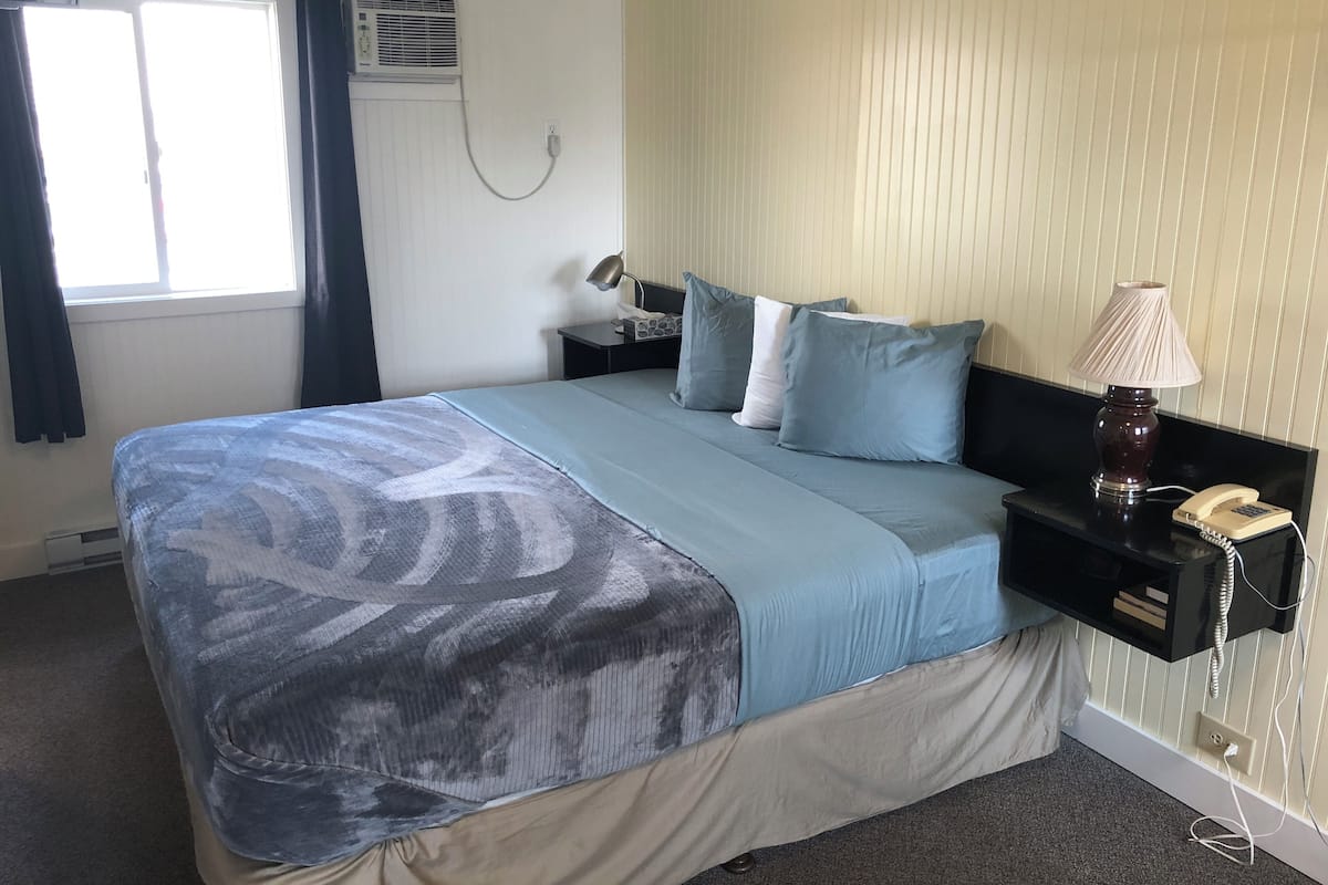 Quarto de hotel com cama, mesa de cabeceira com abajur e telefone, janela com cortinas e aparelho de ar condicionado.