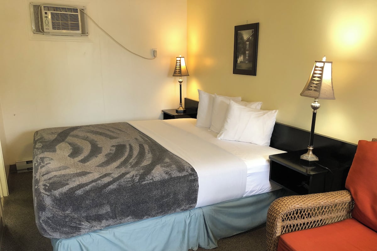 Quarto de hotel com cama, duas luminárias de cabeceira, um quadro na parede e uma cadeira de vime.