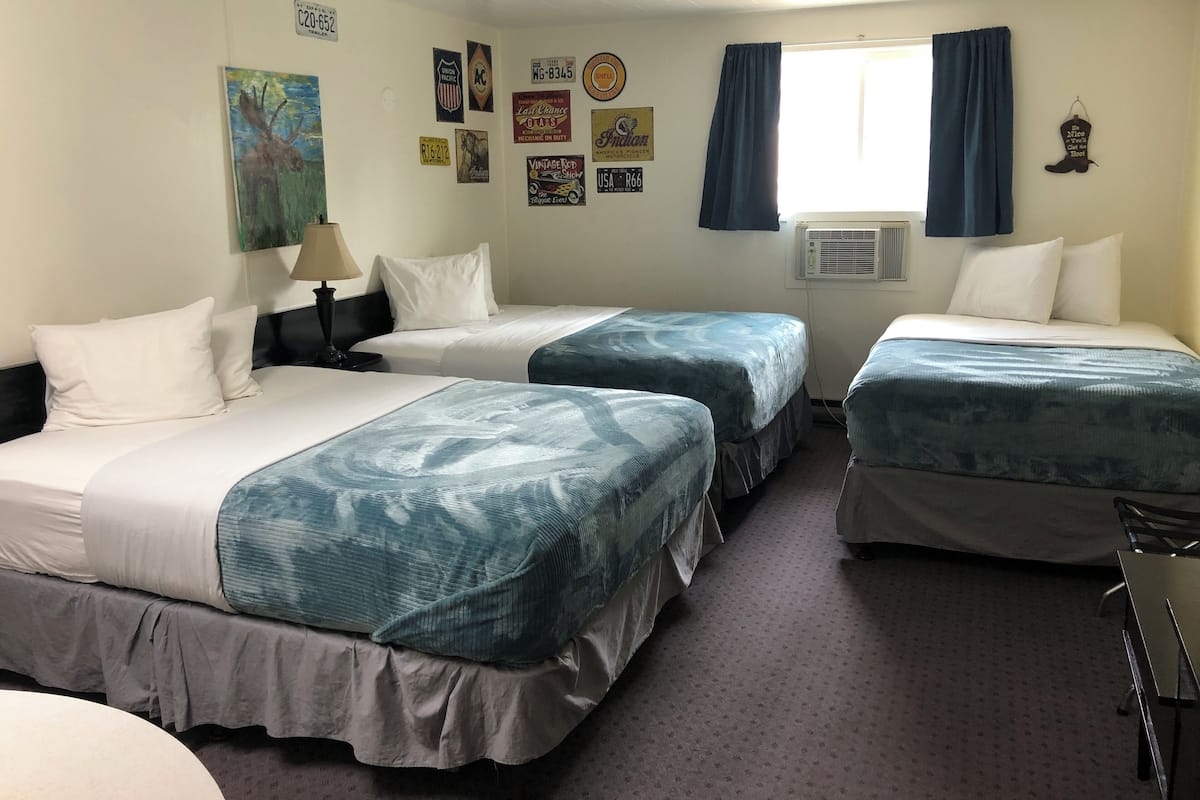 Quarto de hotel com três camas, uma janela com cortinas e várias decorações nas paredes, incluindo placas de carro e uma pintura.