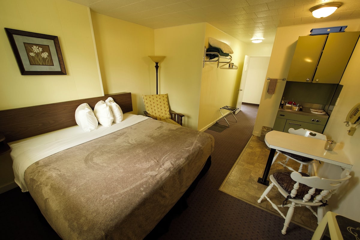 Quarto de hotel com cama, poltrona, escrivaninha e cozinha compacta.