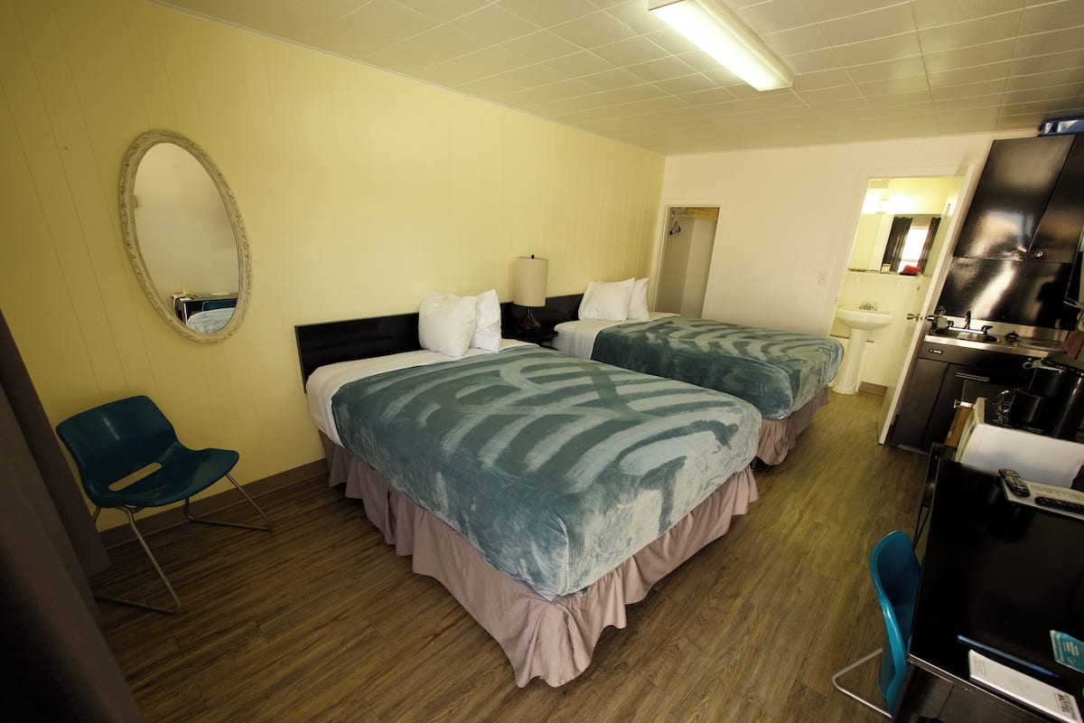Quarto de hotel com duas camas, um espelho, uma cadeira, uma pia e uma cozinha compacta.