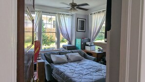 Ein Schlafzimmer mit Bett, Schreibtisch, Stuhl, Deckenventilator und einem Fenster mit Vorhängen.