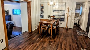 Uma área de refeições com mesa e cadeiras de madeira, um lustre pendente e vista para o exterior através de uma janela.
